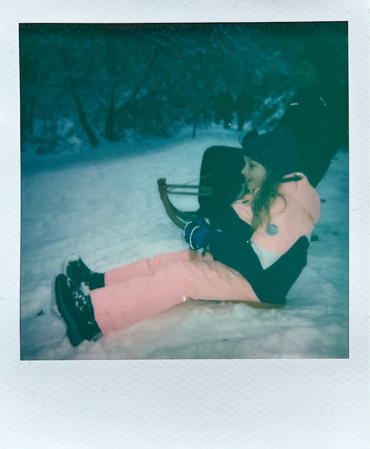 A girl in pink snow clothing on a tiny sledge. A woman on a wooden sledge behind here. Snow and trees all around.