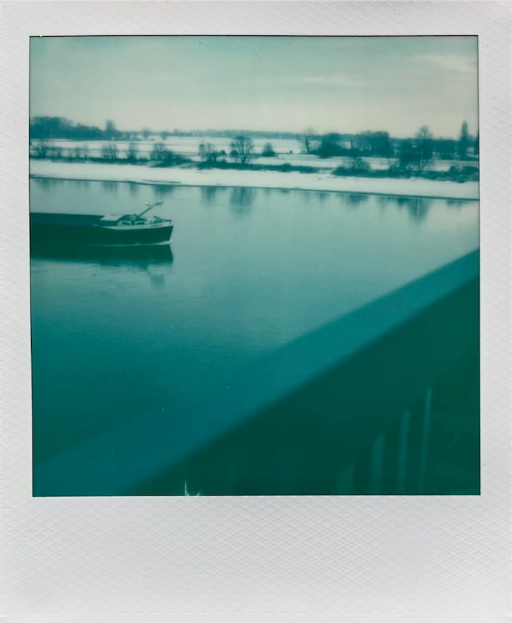 The river Rhine shot from a bridge, which you see a piece of railing in the bottom right. You see the snow covered shore and the horizon behind that in the upper third of the image. The front half of a ship is coming in from the left.