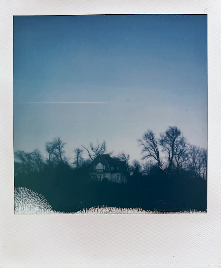 An old house on a hill surrounded by leafless trees. Above is a
      blue sky with a contrail coming in from the left, ending right above the
      house.