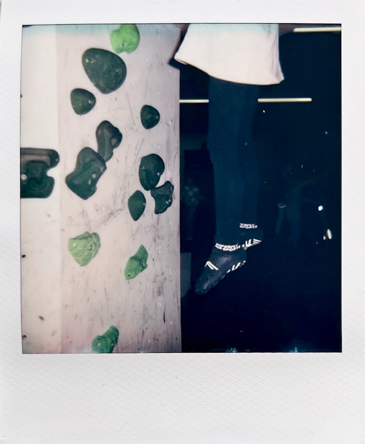 A white boulder wall with green grips on the left side. 50cm
      from the wall to the right, the lower half of a child is hanging
      supposedly in mid-air.