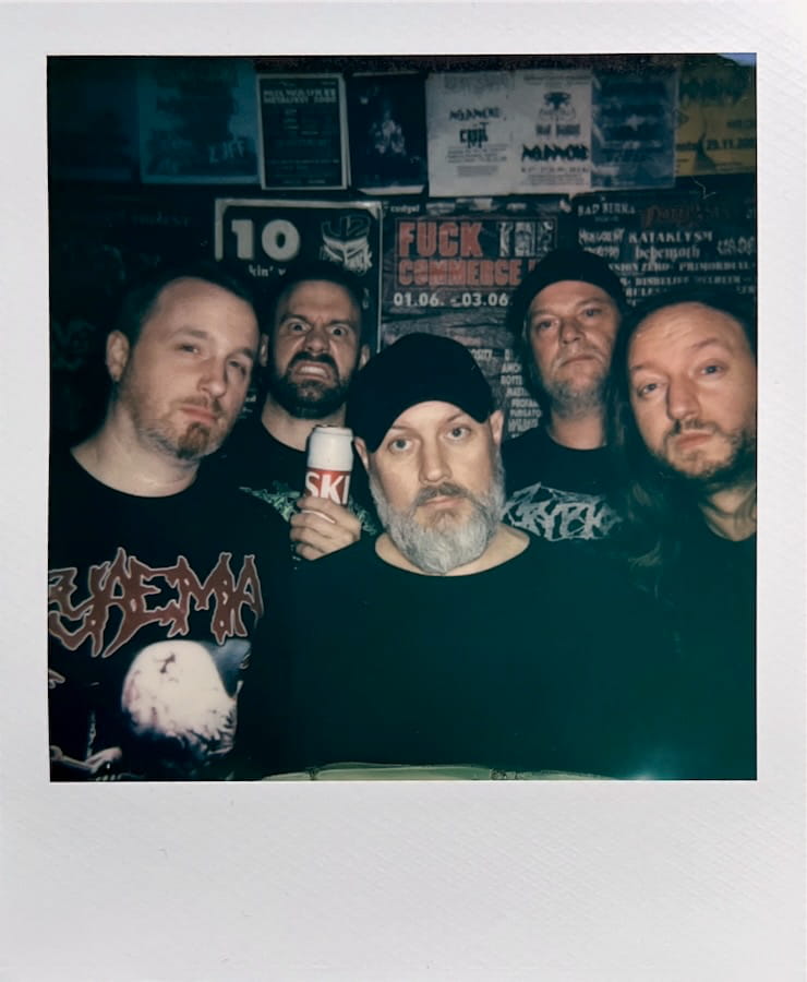 The faces and upper bodies of five men looking into the camera.
      In the background a wall tighly covered in concert posters.