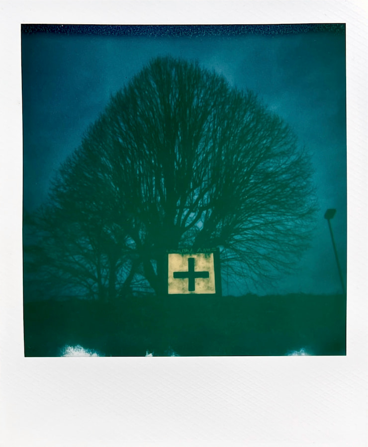 Overall dark (blue) image. The sky in the upper 3/4ths, the
      ground below. A large leafless tree fills the frame. A stone square with a
      cross on it stands on the ground in front of the much large tree.