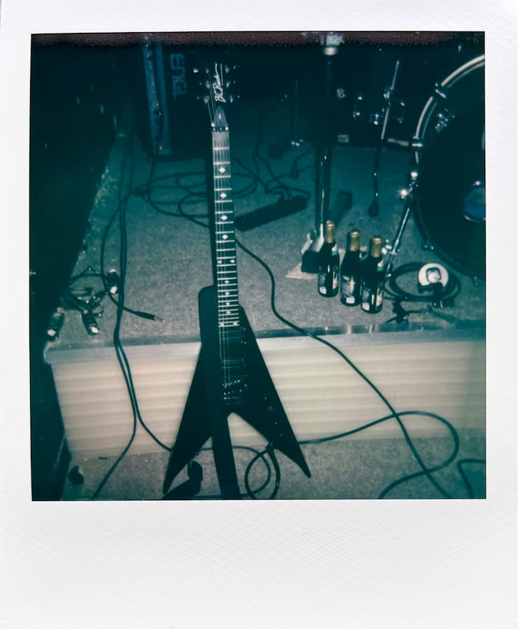 A King-V shaped B.C. Rich guitar leaned against a high step.
      Lots of cables lying around. 3 bottles of beer on the step, as well as a
      small part of a drumkit and an amp head in the background.