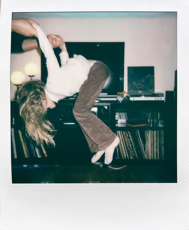 A child mid-air from the side. On the left two adult hands
      holding the child's hands. In the background a cupboard with a TV and
      record player.