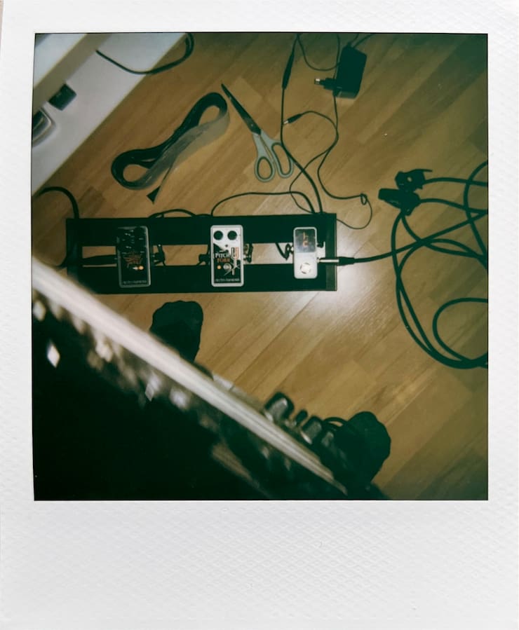 A single-row guitar pedalboard with three pedal on it. Shot from
      above. Cable and other stuff lying around it on the wooden floor.