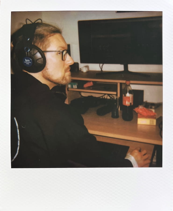A man with headphones on sitting in front of a computer, looking
      back at the camera.