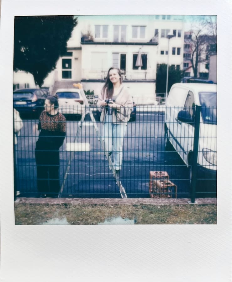 A woman on a latter behind a fence with a camera in hand,
      looking into the camera.