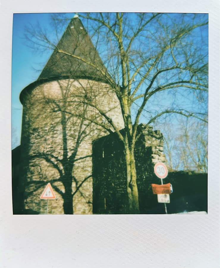 A medieval tower and part of a wall in sunshine with a tree and
      its shadows.