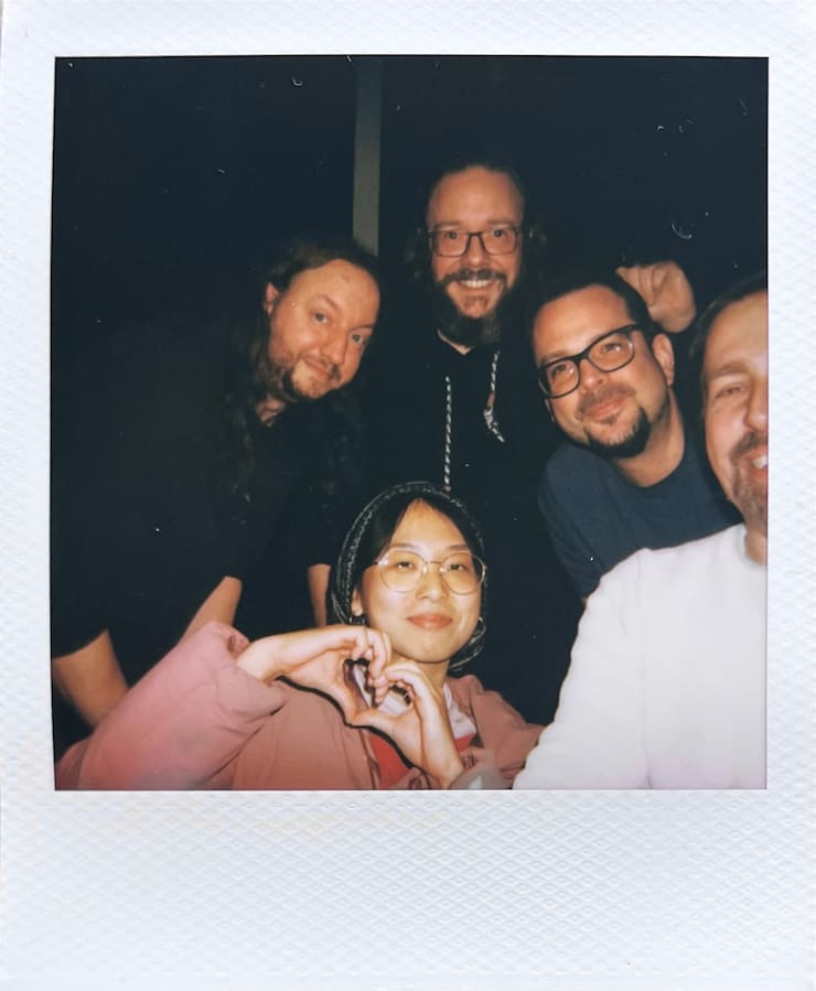 Three and a half men and one woman looking into the camera
      smiling in front of a black background.