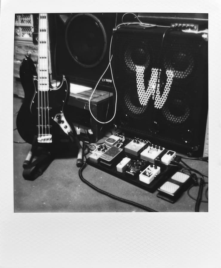 A black & white photo of a Jazz bass, a bass cabinet and a
      pedalboard.