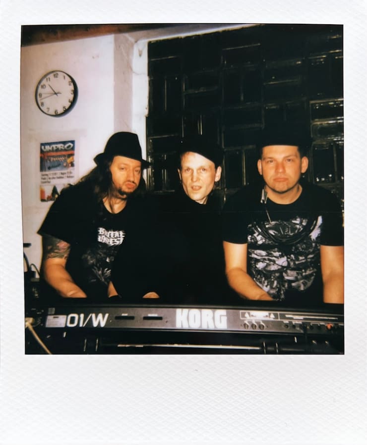 The three men of Forkalyst playing a keyboard. All have hats
      on.