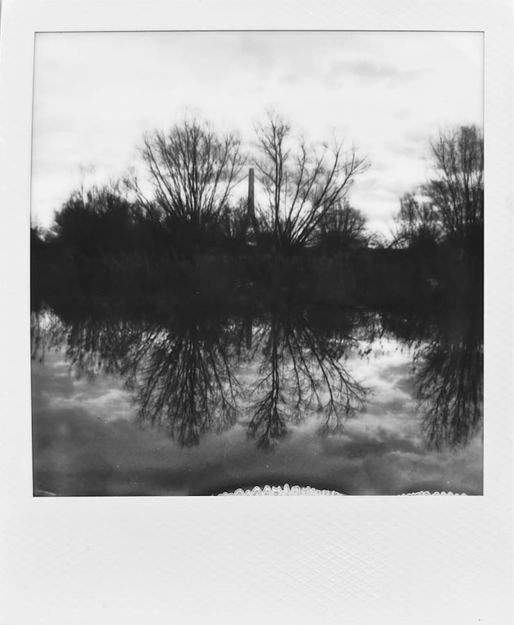 A pond reflecting the leafless trees standing behind it. A
      bridge in the background.