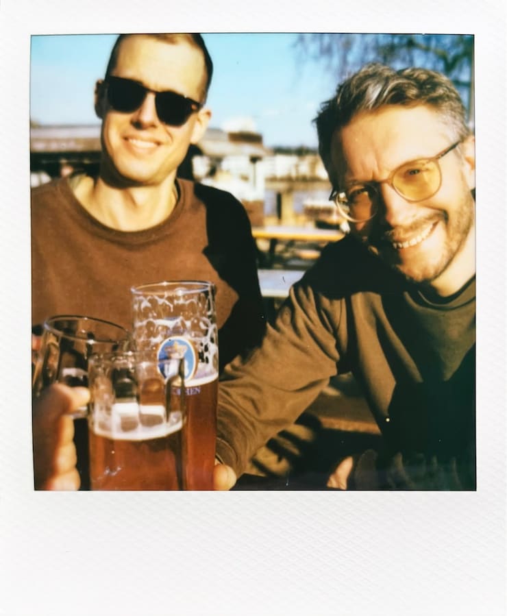 Two men sitting in the sun, looking into the camera and
      cheers-ing with the photographer with glasses of beer.