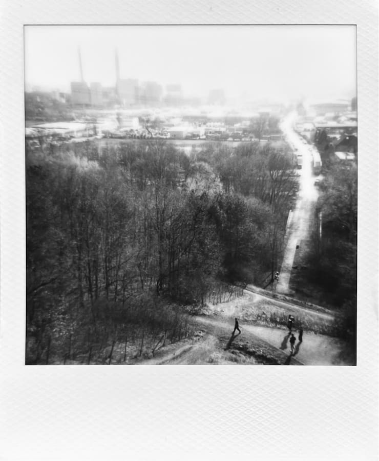 View from higher up over trees, a street and a factory with 2
      chimneys in the sun. Silhouettes of people walking in the
      foreground.