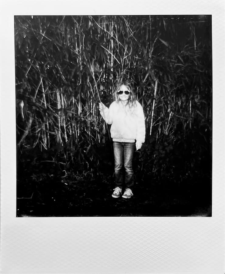 A girl with sunglasses in front of bamboo bush, in a very
      contrasty and rather dark black and white photo.