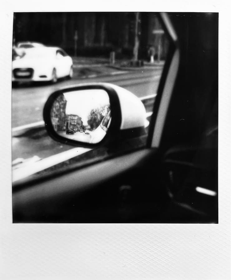 Black and white image from a car to the outside. Cars in the
      rain outside and building in the rear view mirror.