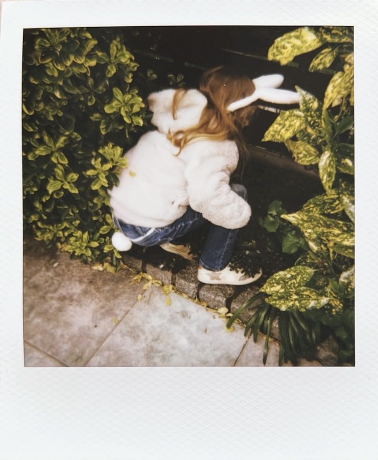 A kid with a bunny decoration searching for Easter eggs amongst garden plants.