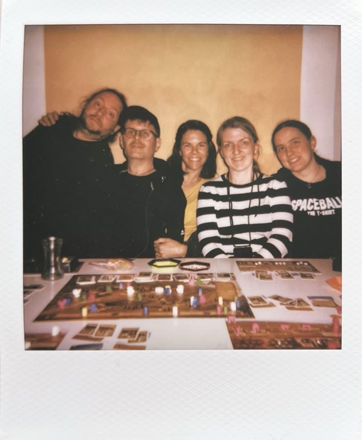 Five people sitting behind a table with a board game on it.