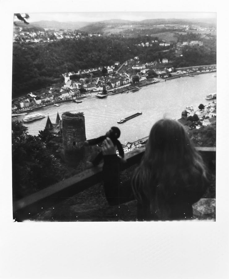 A view of the Rhine from a hillside with a castle and a child in the foreground and a little town in the background.