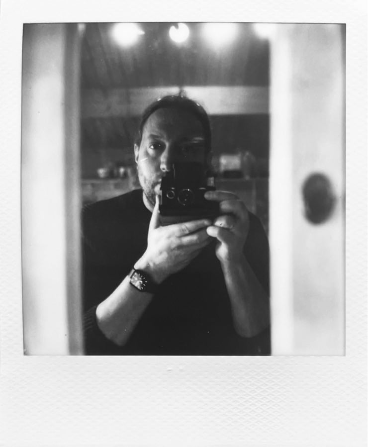 Me and my SX-70 partially visible in a narrow kitchen mirror inside a wooden hut.