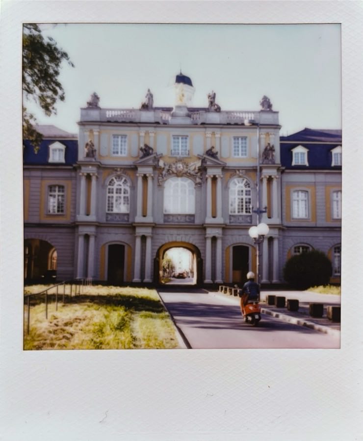 An old grandiose building with a narrow street going through it. A person with a red scooter driving towards it.