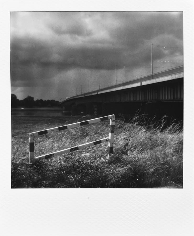 A small barrier in tall grass. A bridge over a river in the background. On the other side are dark clouds.