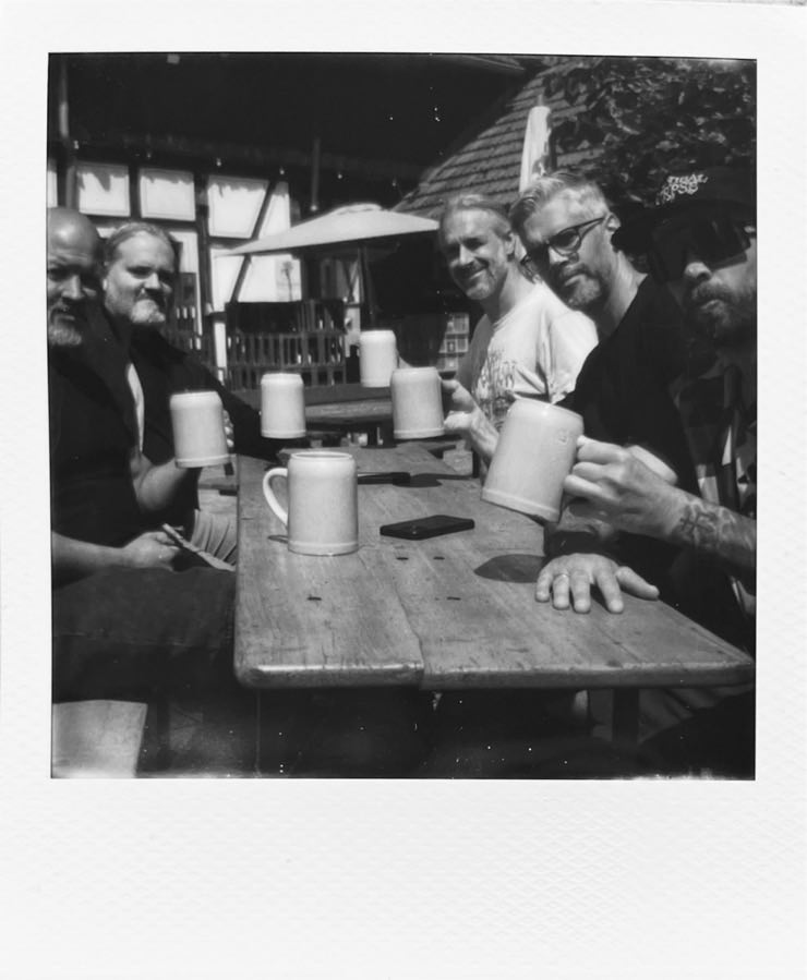 A group of five men sitting at a table in the sun, all having a glass of beer in hand.
