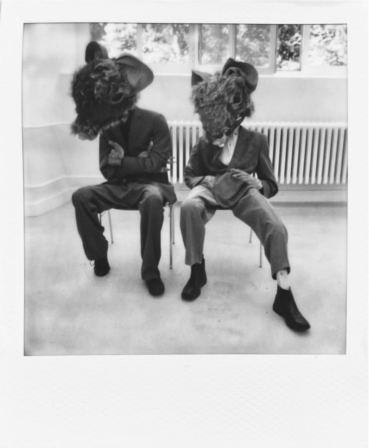 Two seemingly human bodies with giant mice heads sitting on chairs.