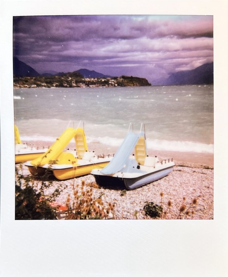A blue and a yellow pedalo on a pebble beach in the sun. The lake in front of dark clouds and mountains.