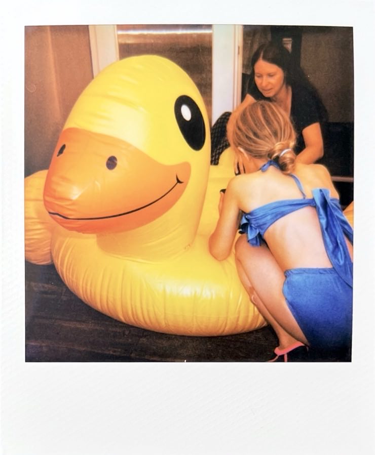 A huge inflatable duck on a porch, a child in swimwear and a woman kneeling beside it.