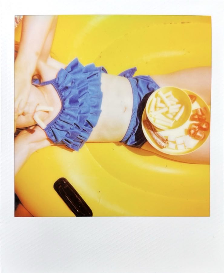 A child on a partly seen yellow inflatable duck, photographed from above. A snack plate is seen and Pasta is used as teeth.