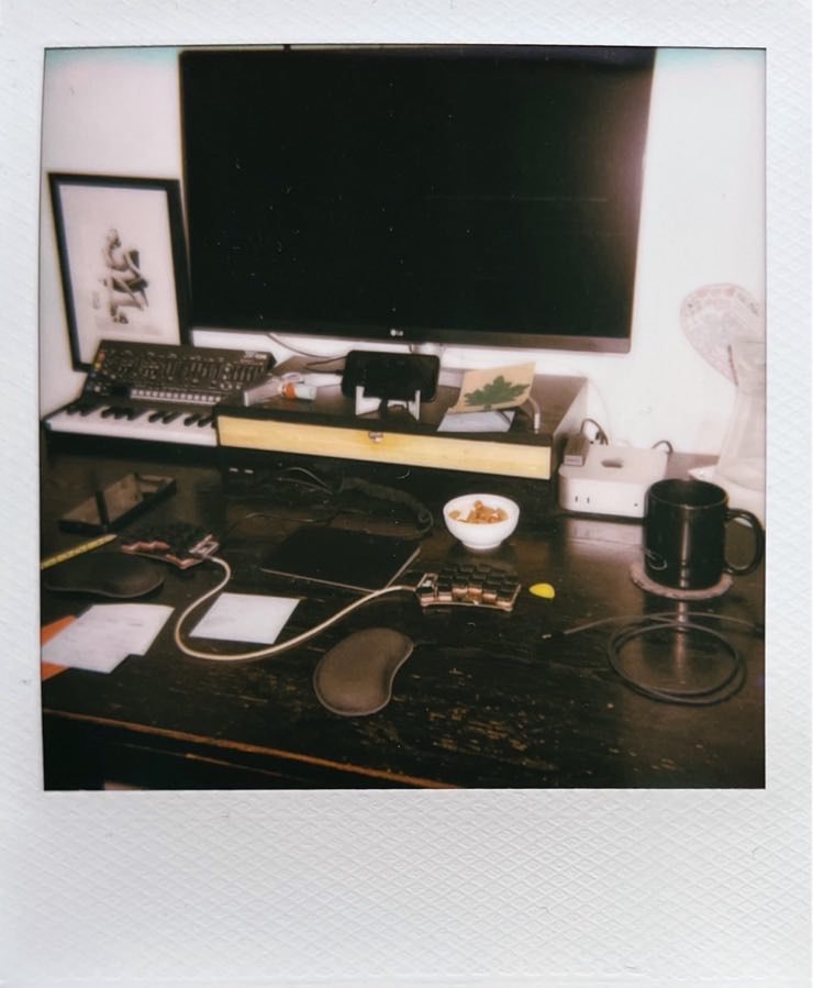 My black desk with a Mac mini, a 27inch screen a split keyboard and a trackpad. A small synth in the background. Some peanuts. A coffee mug.
